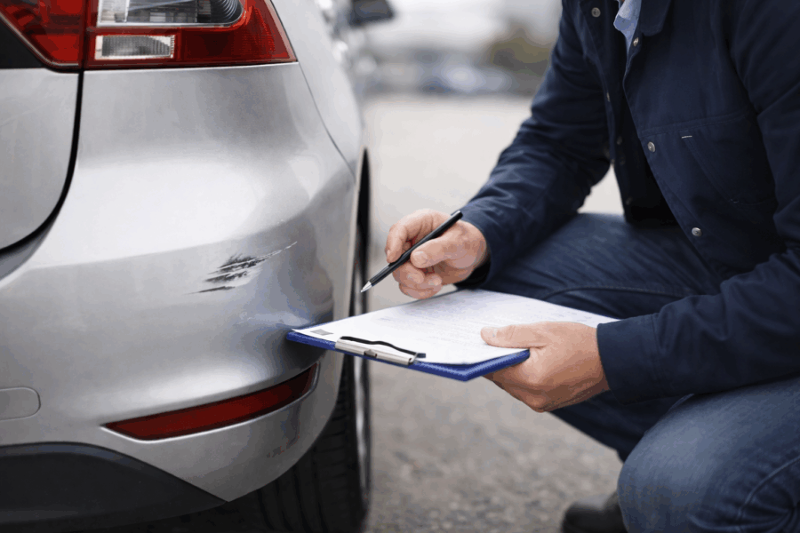 Unabhängiger Kfz-Sachverständiger begutachtet einen kleinen Schaden am hinteren Stoßfänger eines modernen Fahrzeugs und dokumentiert Kratzer und Delle mit Klemmbrett.